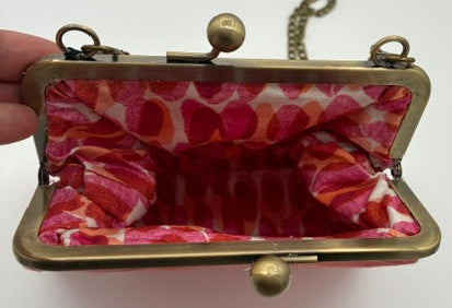 A small kiss-lock handbag with a brass-colored metal frame and a pink and red patterned fabric lining, held open by a hand against a plain white background.