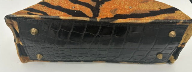 A handbag shown from the bottom front view, featuring a tiger-stripe patterned upper section in orange, black, and tan, combined with a glossy black crocodile-embossed lower section and gold-tone studs.