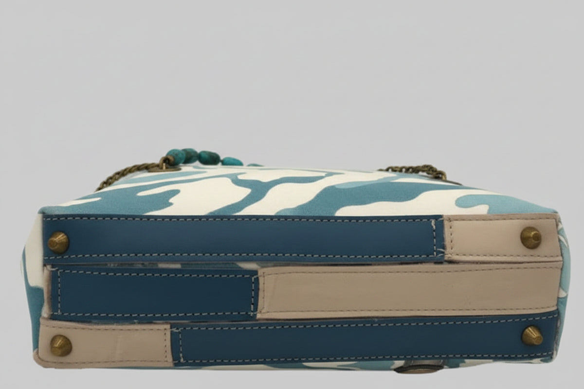 A rectangular patchwork handbag shown from the bottom side, featuring blue leather panels, beige leather trim, brass studs, and a blue-and-white camouflage fabric body with a gold chain strap.