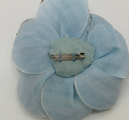 A light blue fabric flower brooch with a textured light blue center and a silver safety pin clasp, shown on a plain white background.