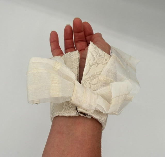 A hand and wrist wrapped in off-white sheer lace and fabric with a bow detail, shown against a plain light background.