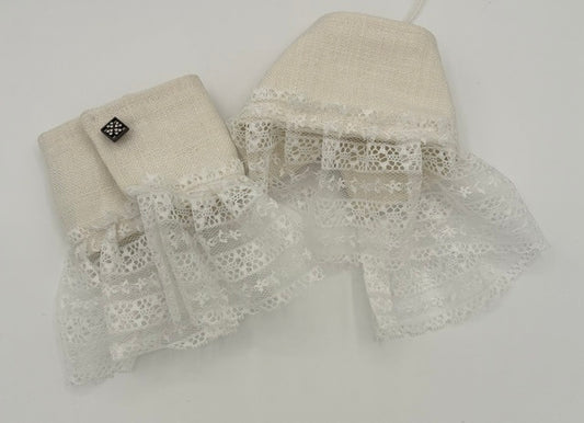A pair of cream-colored linen baby booties with white lace trim and a small black embroidered logo patch, displayed on a light gray background.