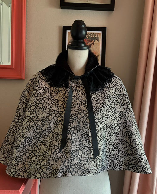 A black and SILVER FORMAL  floral WEAVE cape with a black ruffled collar and ribbon tie, UNDER A BLACK HIDE COLLAR  displayed on a mannequin torso in an indoor room setting.