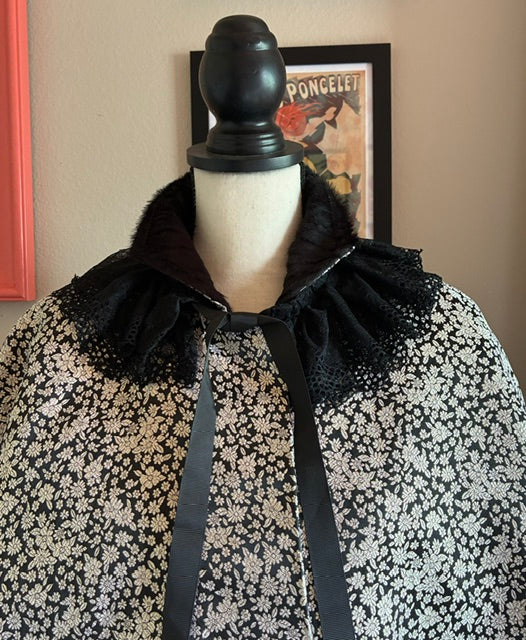 A black and SILVER floral-patterned cape with a black faux-fur collar and a wide black ribbon LACE COLLAR  displayed on a mannequin torso.