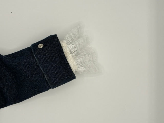 A close-up of a dark navy blue denim sleeve with a white lace ruffle cuff and a single silver button, set against a plain light gray background.