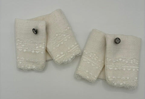 white colored fabric cuffs with white lace and scalloped edges, each featuring a small pewter  metal button, arranged on a plain light gray background.