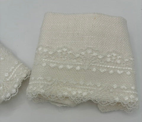 close up of a white lace cuff on a white woven fabric trim or ribbon with a decorative embroidered pattern and scalloped lace edge, folded on a plain light gray background.