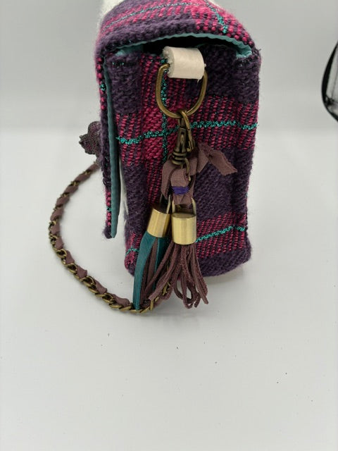 A small woven clutch or pouch in a purple and magenta plaid pattern with teal stripes, featuring a brown leather tassel, gold-tone metal accents, and a braided chain strap, photographed on a plain white surface.