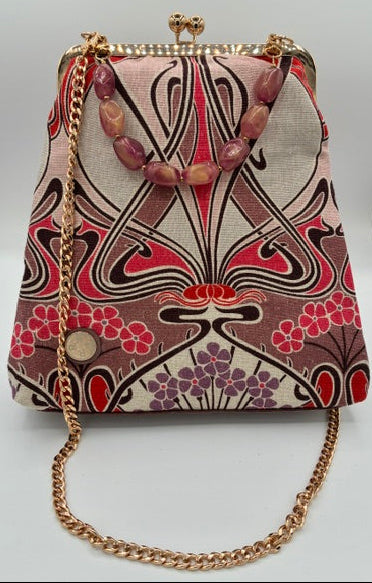 A small handbag with a pink, red, and burgundy abstract art nouveau patterned fabric body, a gold-tone chain strap, and a decorative pink beaded strap detail, shown on a white background.