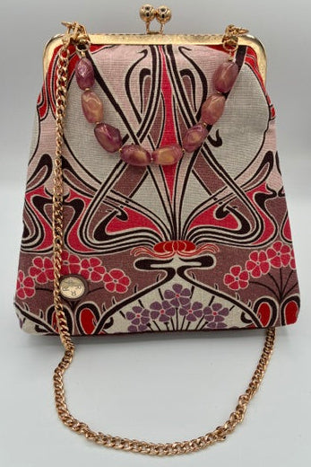 A small handbag with a geometric art deco pattern in pink, red, black, and beige, featuring a gold-tone kiss-lock clasp, a gold-tone chain strap, and a pink stone bead collar.