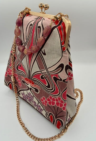 A small handbag with a gold-tone kiss-lock clasp and chain strap, featuring a pink, red, and beige abstract patterned fabric, displayed on a white background.