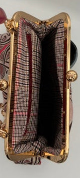 A close-up of a hand holding a small gold-tone kiss-lock handbag with a pink and red plaid patterned fabric lining and a matching pink beaded wristlet strap, set against a white background.