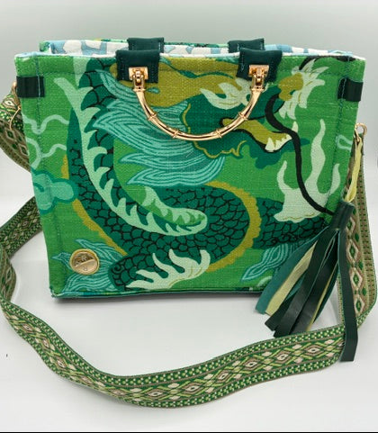 A green handbag with a dragon print, featuring dark green leather handles, a gold-tone metal ring, and a wide patterned green and white shoulder strap, displayed on a white surface.