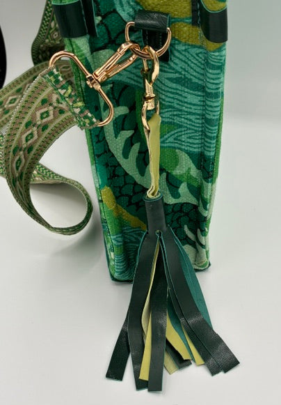 Close-up of a green patterned handbag with a matching patterned shoulder strap and a black leather tassel keychain with gold-tone hardware, set against a plain white background.