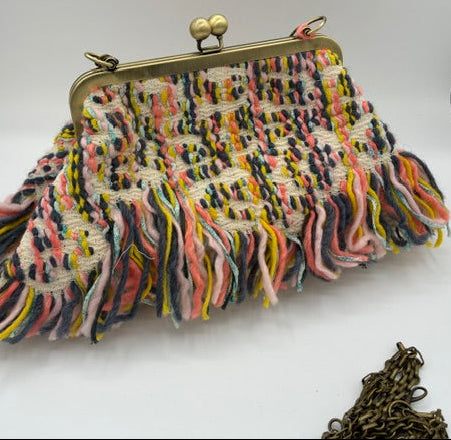 A multicolor woven clutch purse with a fringed tassel edge, antique brass frame, and kiss-lock clasp, displayed on a white surface.