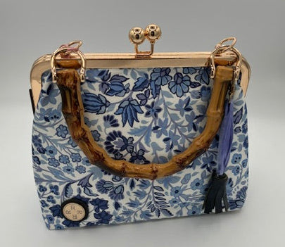 A small handbag with a blue and white floral pattern, a curved brown handle, and gold-tone metal hardware, photographed on a plain light background.