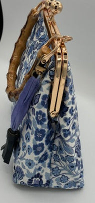 A small handbag with a blue and white floral pattern, a purple tassel, and a gold-tone metal clasp, shown against a plain light gray background.