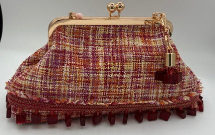 A small tweed kiss-lock handbag in a red, orange, yellow, pink and beige plaid pattern with a fringed bottom and gold-tone metal hardware, displayed on a white surface.