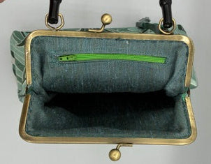 A vintage-style clutch handbag with a muted green silk interior, a gold-tone metal frame, and a black bamboo handle, held open by a person's hand against a plain white background.