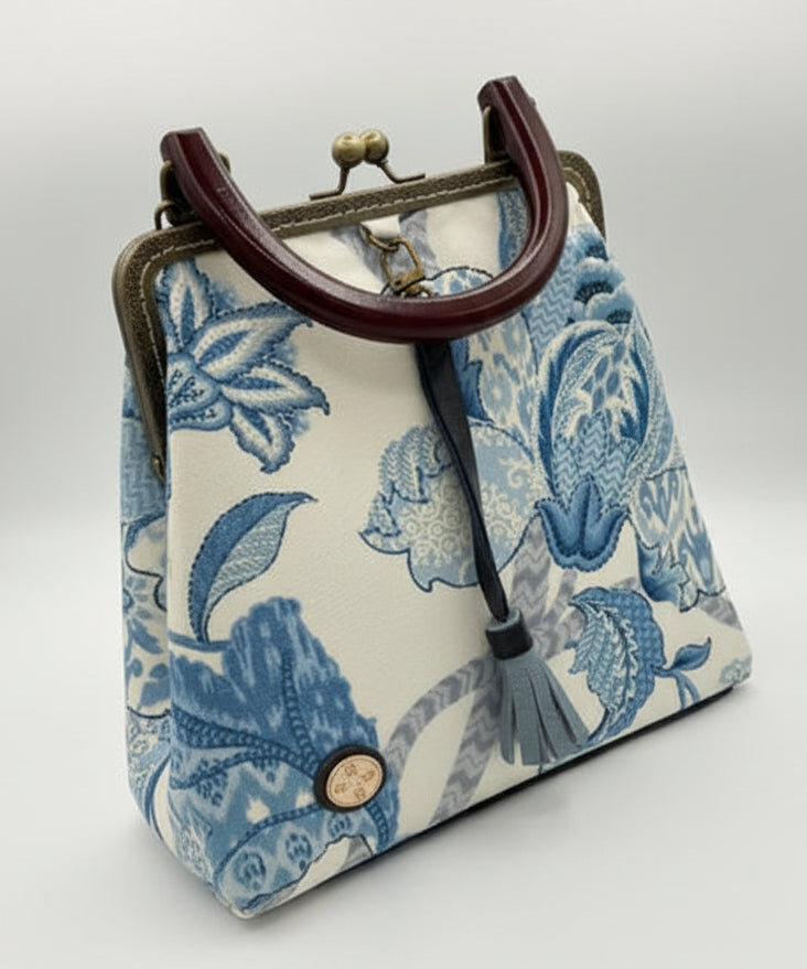 A small fabric handbag with a blue floral toile pattern on an off-white background, featuring a dark brown leather handle, brass-tone clasp, and a gray tassel. A hand is pointing at the bag.