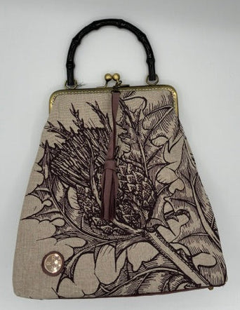 A small handbag with a woven beige body featuring a dark purple floral and leaf pattern, a black acrylic bamboo handle, and a gold-tone metal frame.