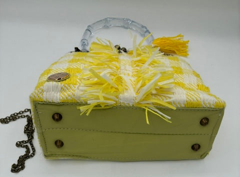 A structured handbag with a yellow and white plaid patterned top, yellow fringe trim, a clear plastic top handle, and a light olive green base. It features dark metal studs on the sides and a dark chain strap, photographed on a plain white background.