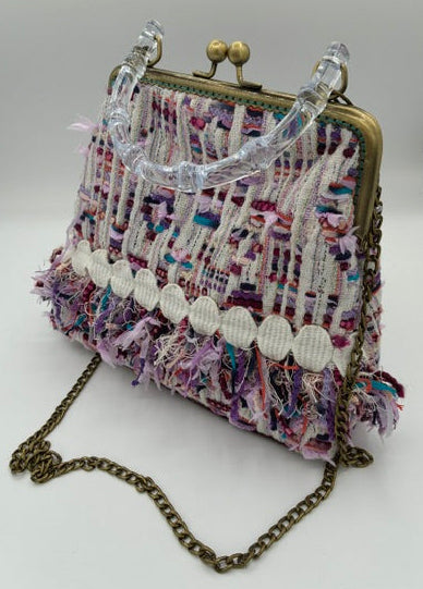 A small woven clutch handbag with a multicolor tweed pattern in purple, pink, and white, featuring a clear plastic handle, antique gold-tone metal frame, and a gold-tone chain strap.