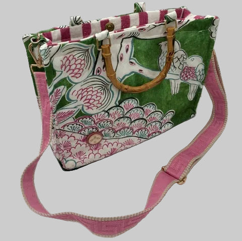 A fabric shoulder bag with a green, pink, and white floral print, featuring a pink adjustable strap, a brown faux-bamboo handle, and a pink-and-white striped side panel.
