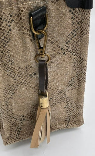 A close-up of a beige snakeskin-patterned tote bag with black leather trim, a gold-tone clasp, and a matching leather tassel keychain, held by a hand against a white background.