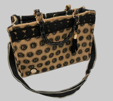 A structured tote bag with a tan and black monogram pattern, featuring black lace trim along the top, black leather handles, a black tassel charm, and a black-and-tan shoulder strap.