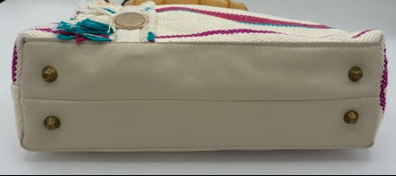 A cream-colored shoulder bag shown from the bottom, featuring brass studs on the base, a colorful woven strap, and a bamboo handle, photographed against a white background with black side panels.