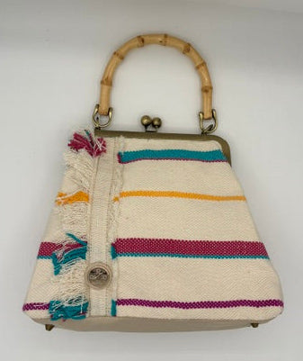 A small woven handbag with a bamboo handle, featuring horizontal stripes in teal, yellow, magenta, and orange on an off-white base. It has a button closure with frayed tassels on the front and a metal frame clasp at the top.