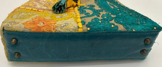 A vintage patchwork handbag with a teal leather base, a yellow fabric handle, and a floral embroidered top panel in beige, yellow, and orange.