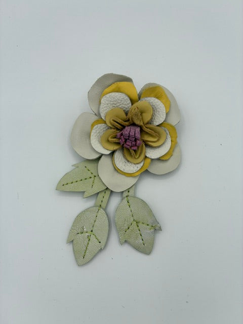 A handmade leather flower embellishment with layered petals in olive green, cream, and a purple center, accompanied by two stitched sage green leaves on a plain white background.