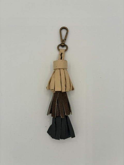 A three-tiered leather tassel keychain with a brass carabiner, featuring light beige, dark brown, and black leather layers, shown against a plain light gray background.
