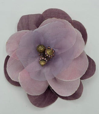 A fabric flower hair accessory in shades of dusty purple and lavender, featuring a textured center with small gold and brown beads, set against a plain white background.