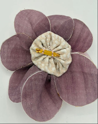 A fabric flower brooch with muted purple petals and a textured beige center, featuring a gold safety pin clasp.