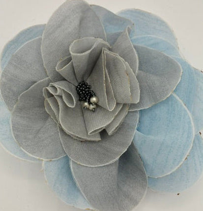 A fabric flower accessory with layered light blue and gray petals, featuring a small black beaded center with two pearl-like accents, shown on a plain light background.